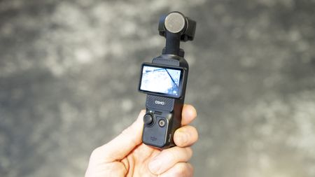 DJI Osmo Pocket 3 vlogging camera in user's hand, grey floor in background